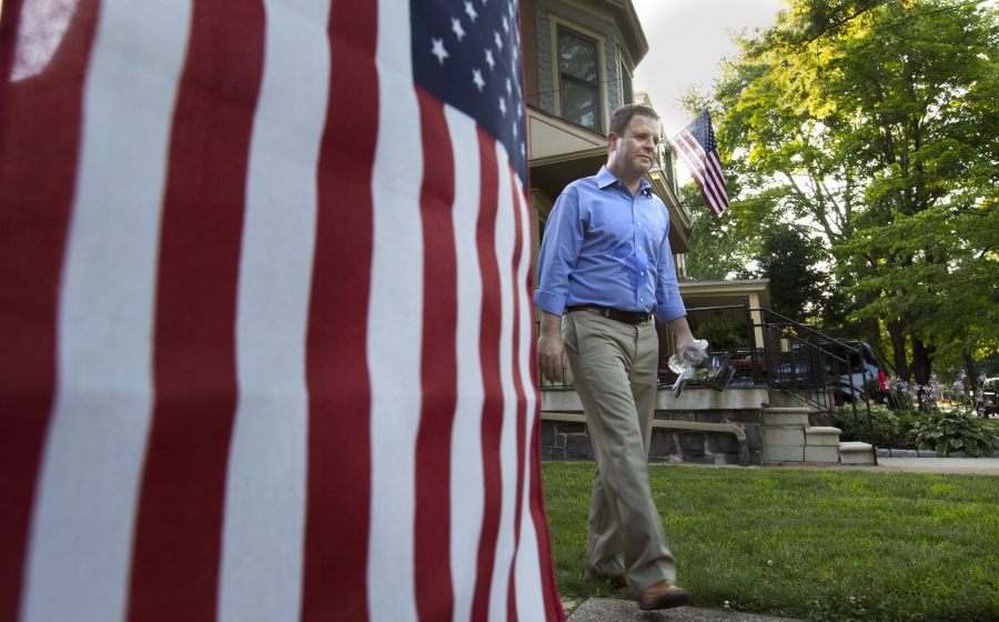 a campaigning politician in a blue shirt and khaki pants walks down a driveway past an American flag